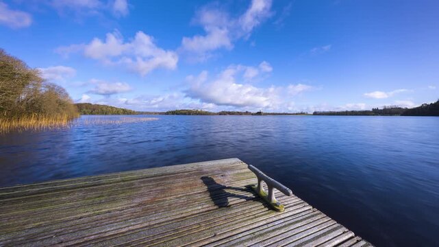 Time lapse of a timber lake pier in the foreground and castle ruin island with forest in distance on a cloudy sunny day at Lough Key in county Roscommon in Ireland.