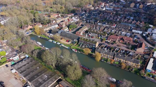 Stanstead Abbotts drone reverse over the River Lea and High Street, revealing canalside homes, moored narrowboats, winter trees and town rooftops in Hertfordshire, England.
