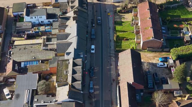 Stanstead Abbots high street drone flyover toward the railway station, showing shop shadows, light traffic, parked cars, and road markings in a small English town.