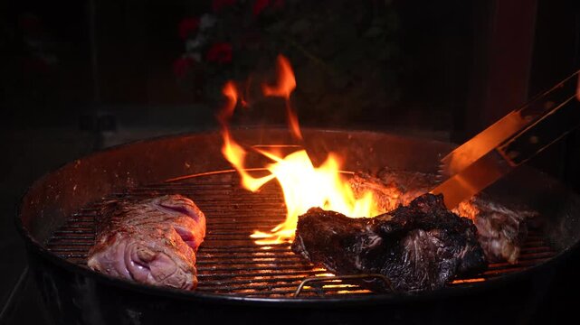 A bbq grill with 3 pieace of meat being cooked. A large flames dies down after having burnt one of the pieces of meat. A man with bbq tongs checks and moves the meat.