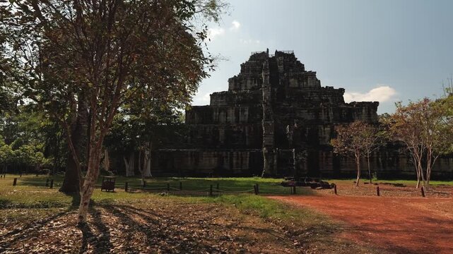 Walking toward Koh Ker pyramid temple ruins along red dirt path, framed by trees under blue sky