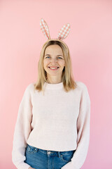 Pretty middle-aged blonde woman with bunny ears decoration standing in front of pink background