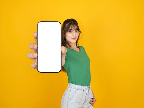 Cheerful young woman showing smartphone with blank white screen for mockup, looking at camera and smiling. Professional presentation of  mobile app or digital service against vibrant yellow background