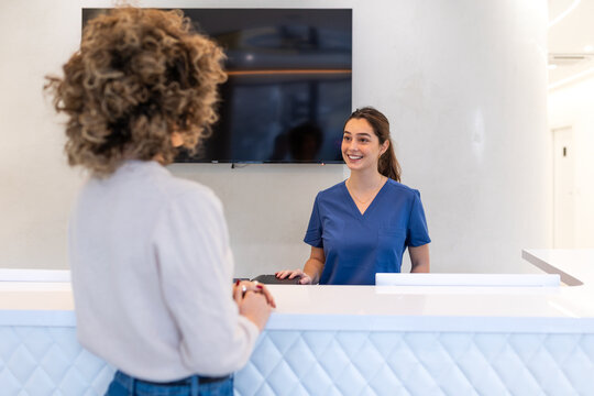 Smiling receptionist in blue scrubs greeting patient at clinic front desk. Professional medical staff at reception area in modern hospital. Healthcare administration and service concept image.