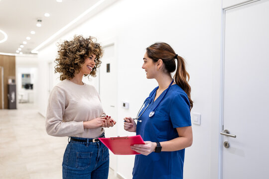 Friendly nurse in blue scrubs consulting young patient in clinic hallway. Professional medical staff holding clipboard talking to woman in hospital corridor. Healthcare and insurance concept image.