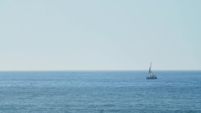 A small sailing boat is sailing in the open ocean on a bright sunny summer day against a blue cloudless sky. A tiny sailing vessel drifts through the vast open ocean.