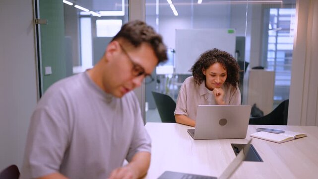Man with glasses concentrates on laptop and handwritten notes while female coworker works in background. Clean open office space highlights cooperation and business productivity.