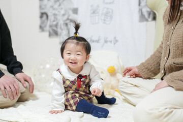 Naklejka premium A baby girl is sitting on a bed with her hands on her face. She is smiling and looking at the camera. The room has a white background and a few other objects, including a stuffed animal and a balloon