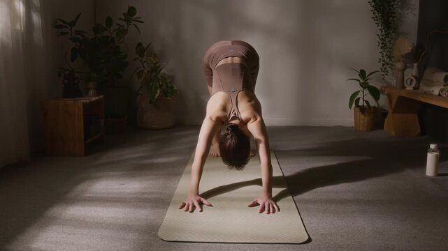 Wide frontal view of motivated young Caucasian woman doing routine yoga practice on mat at home, assuming downward dog or adho mukha svanasana, with focus on correct posture