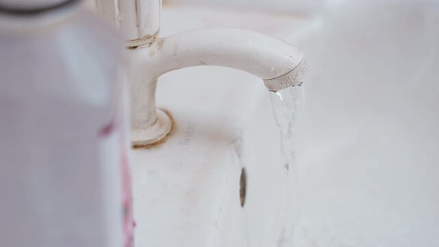 White sink faucet dripping, aged porcelain basin with soap bottle, slow water flow, mineral stains and rust around fixture, intimate closeup of domestic maintenance and routine bathroom chore