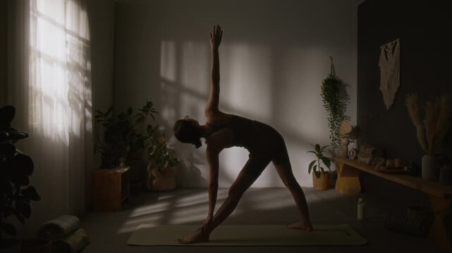 Full shot of female yoga enthusiast doing trikonasana, then changing to high lunge alanasana posture, maintaining concentration and awareness, dark silhouette in dim window light