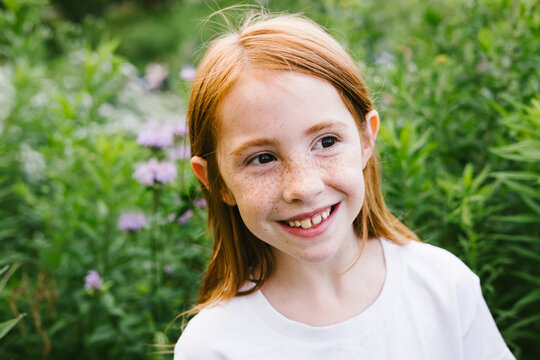 Red haired girl with freckles smiles with field of grass and flowers