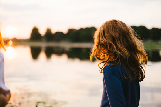 Red haired female child by edge of lake at sunset with trees