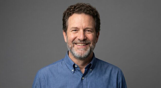Smiling middle-aged man with a beard and curly hair looks directly at the camera, exuding confidence and approachability on a solid gray background