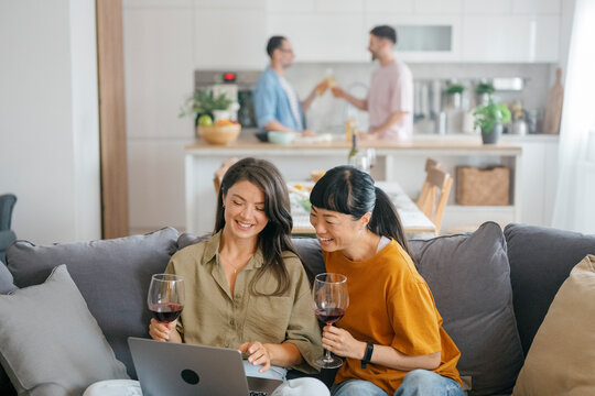 Friends sitting on couch with wine using laptop and laughing at home