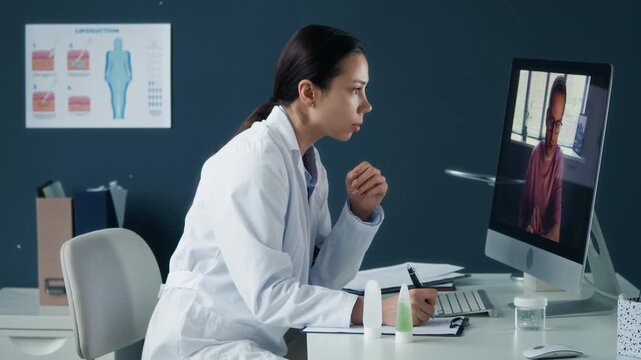 Female dermatologist conducting telemedicine consultation with patient on desktop computer in modern clinical office, eyelevel side view medium shot with medical workspace interior