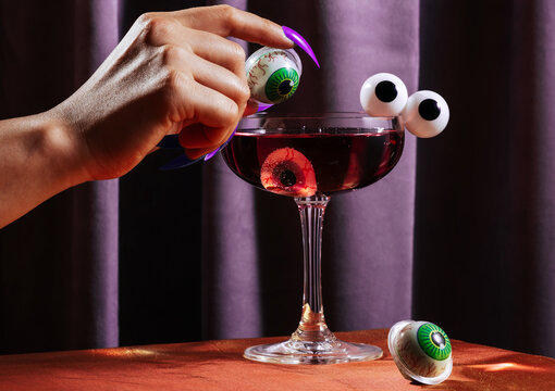 Witch hand with fake nails holding eyeball garnish over purple cocktail