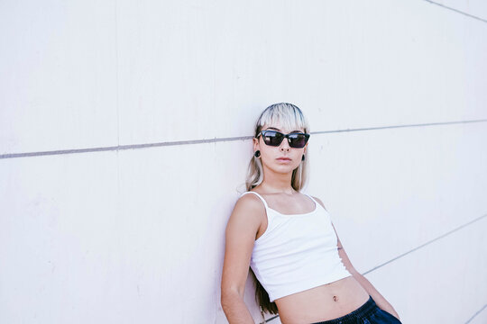 Young woman with attitude in sunglasses and white tank top leaning on urban wall