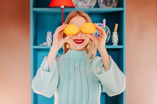 Smiling woman in blue dress holding lemons at eyes in colorful interior