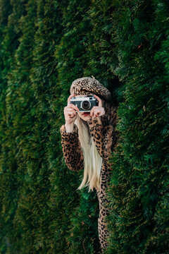 Photographer in leopard print stylish jacket hiding in thuja garden taking picture