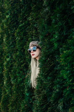Stylish woman with sunglasses and leopard jacket posing by thuja hedge