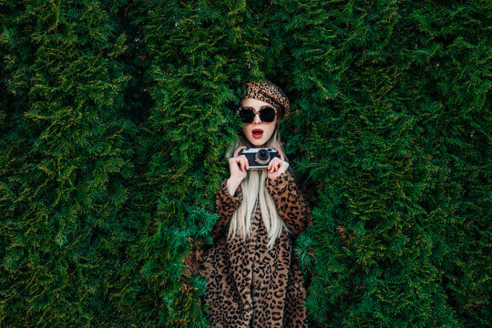 Stylish woman in leopard jacket with camera outdoors in lush thuja garden