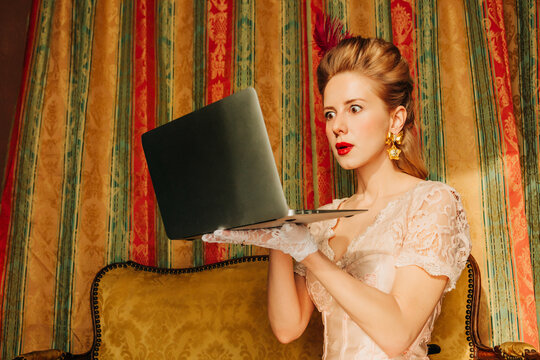 Vintage styled woman in 18th century dress shocked using laptop indoors