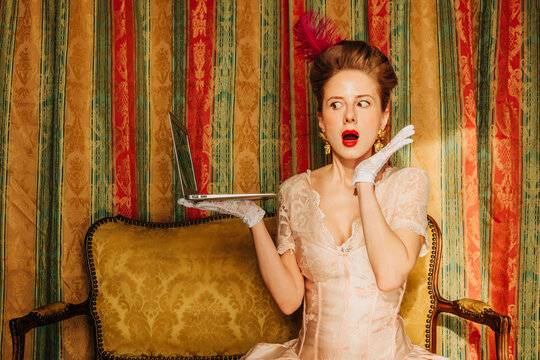 Surprised woman in Rococo vintage dress using laptop on vintage sofa