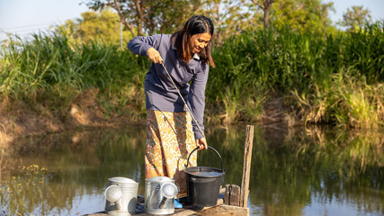 A woman is collecting water from a river using a bucket. She stands on a wooden platform with two containers nearby. The surroundings show greenery and trees © Happy Photo
