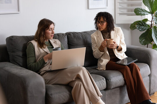Diverse women colleagues sitting on sofa in lounge, using laptop, tablet and mugs in blazer, vest