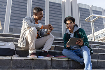 Diverse male friends sitting on concrete steps at campus, using tablet with stylus and notebooks