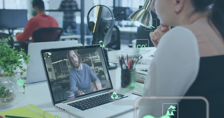 Watching woman wearing layered top, leaning at open-plan desk; laptop, plant, eco icons, copy space