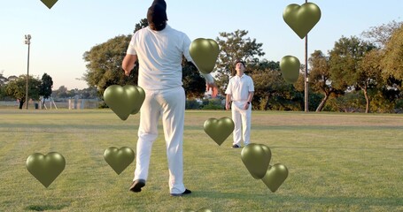 Walking two white-clad cricketers approaching on turf late-afternoon, metallic olive heart balloons