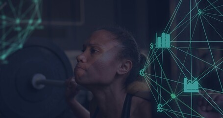 Squatting woman pushing through back squat at gym, wearing dark tank, loaded barbell and teal data