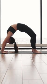 A woman in black activewear performs a backbend yoga pose on a mat in front of a large window with a city view