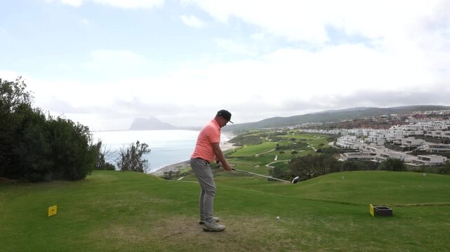 Golfer in pink shirt prepares to swing on La Hacienda golf course, overlooking ocean and coastal landscape with residential buildings
