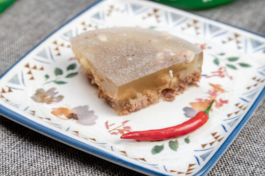 Closeup photograph of a single portion of homemade holodets, a traditional Russian cold meat appetizer (aspic or studen), on a square plate in a green bowl.