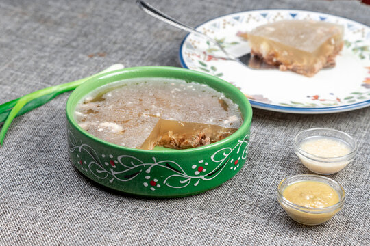 Closeup photograph of a single portion of homemade holodets, a traditional Russian cold meat appetizer (aspic or studen), on a square plate in a green bowl.