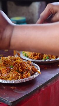 ​Vertical close-up video of a street food vendor serving a hot and spicy Indian noodle dish onto silver foil plates at a local stall
