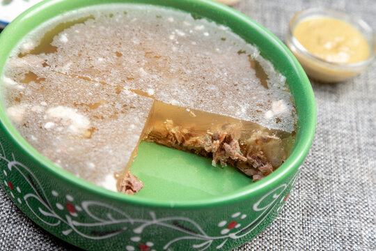 Closeup photograph of a single portion of homemade holodets, a traditional Russian cold meat appetizer (aspic or studen), on a square plate in a green bowl.