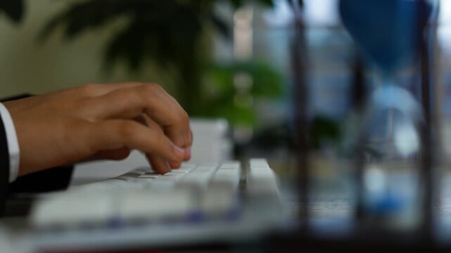 Hands typing on white keyboard with hourglass, Close-up of hands typing on a white keyboard with a blurred office background and hourglass