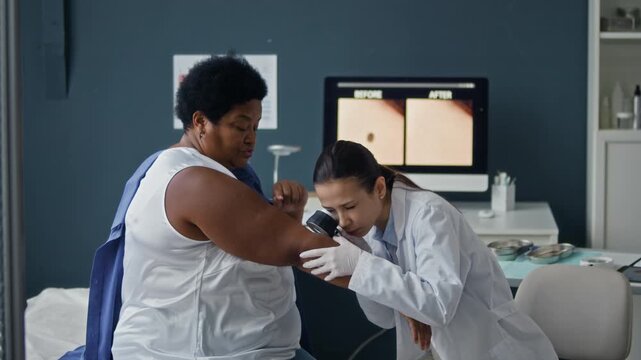 Dermatologist inspecting suspicious mole on African American female patient arm during medical consultation with diagnostic image visible on computer screen in background, side view medium shot