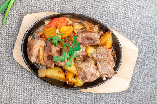 Sizzling cast iron skillet with braised kid meat and roasted potatoes on a wooden board, closeup.