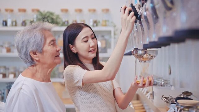 Young Asian woman and her senior mother enjoy shopping together, choosing natural products from a bulk dispenser at a modern zero waste refill station store.