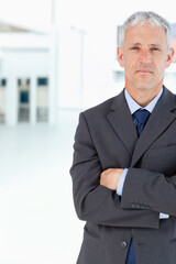 Fototapeta premium Man standing with crossed arms facing forward in modern lobby wearing dark suit, copy space