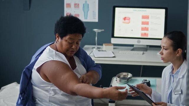 Medium side view eye level shot of young Caucasian female dermatologist providing skin examination as Black female patient presenting inner upper arm during consultation in medical office