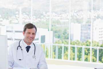 Mature doctor sitting at desk in clinic with computer wearing lab coat and stethoscope, copy space