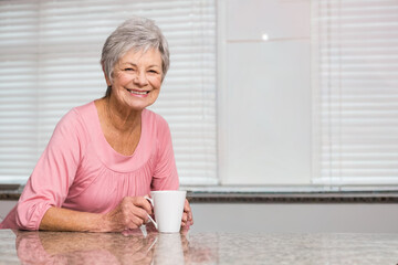 Senior woman leaning on granite counter holding mug in pink by kitchen window blinds, copy space