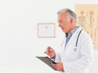 Senior doctor reviewing clipboard in clinic wearing coat and stethoscope, holding pen, copy space