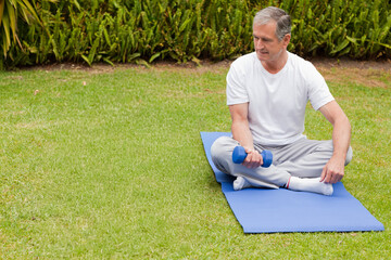Senior man sitting on blue exercise mat on lawn holding small blue dumbbell, copy space